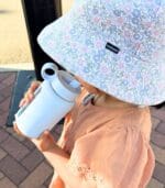 Young child in a floral sun hat drinking outdoors from a white Kaally glass water bottle