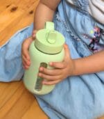 Toddler sitting on a wooden floor holding a sage green Kaally glass water bottle with a straw lid