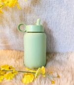 Sage green Kaally glass water bottle resting on a sheepskin rug beside yellow flowers