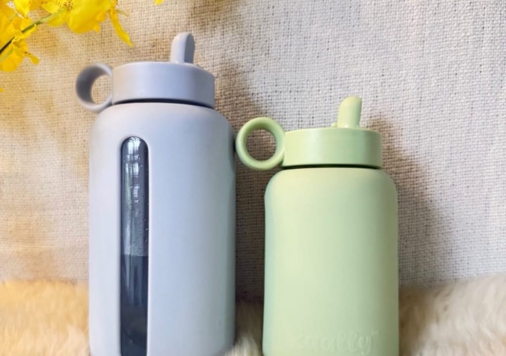 Two Kaally borosilicate glass water bottles with silicone sleeves and straw lids, styled with yellow flowers