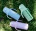 Three pastel KaakaaBaby kids glass water bottles with straw lids on grass, ready for engraved names