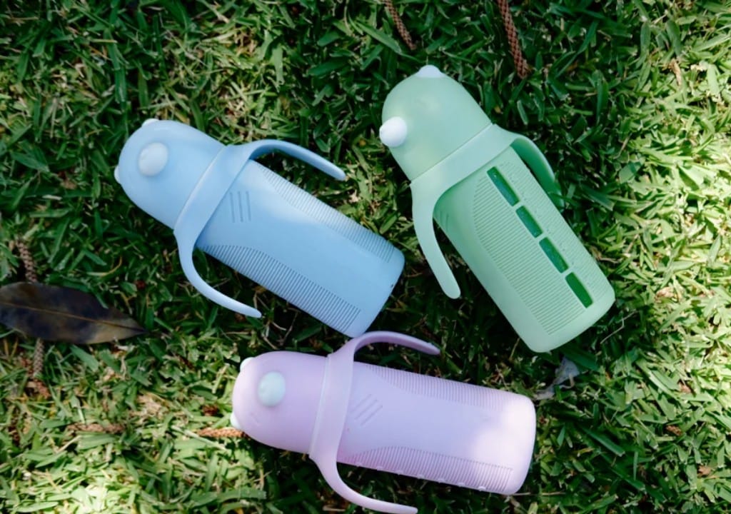Three pastel KaakaaBaby kids glass water bottles with straw lids on grass, ready for engraved names