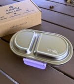 Kaally Snack Box New 2026 Edition in Lilac with its kraft retail box on an outdoor table — two-compartment 18/8 stainless steel snack container with food-grade silicone seal