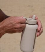 Adult hands holding the Kaally 800mL glass water bottle in Almond with the sports lid fitted, on a beach